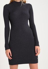 Robe en maille côtelée gris foncé à manches longues, col monté, et silhouette cintrée. Présente un détail de logo discret à l'ourlet.