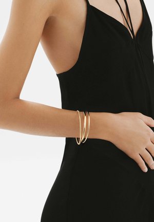 Brazaletes de oro en una muñeca, variando en grosor, con un acabado liso y brillante. Prenda negra con tirantes delgados en el fondo.