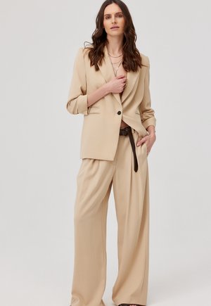 Kobieta z długimi brązowymi włosami, ubrana w beżową marynarkę i szerokie spodnie, stojąca z jedną ręką na piersi, a drugą w kieszeni.