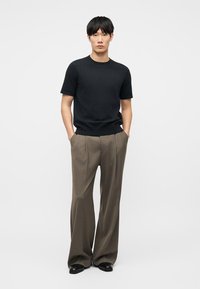 Νεαρός άνδρας στέκεται με τα χέρια στις τσέπες, φοράει μαύρο πουκάμισο με κοντά μανίκια, καφέ παντελόνι φαρδύ και μαύρα παπούτσια, μπροστά σε απλό φόντο.