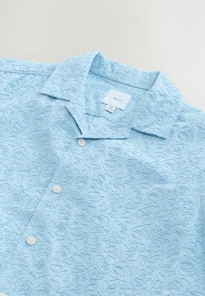 Lichtblauw kortarm shirt met een algehele textuur met bloempatroon, twee knopen en een puntige kraag. Merklabel zichtbaar aan de binnenkant.
