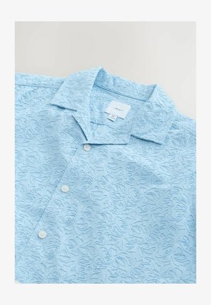 Chemise à manches courtes bleu clair avec un motif floral texturé sur l'ensemble, deux boutons et un col pointu. Étiquette de marque visible à l'intérieur.