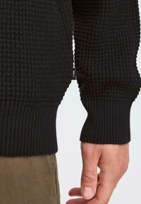 Schwarzer strukturierter Pullover mit Waffelstrickmuster, gerippten Bündchen und einem sichtbaren Markenetikett an der Naht. Hand ruht nahe dem Saum.