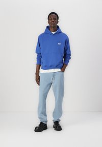 Blaues Kapuzenoberteil mit einer Vordertasche und weißem Logo, kombiniert mit hellblauen Jeans und schwarzen klobigen Schuhen. Glatte Textur, lässige Passform.