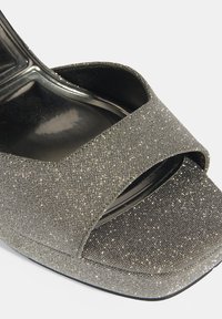 Open-toe wigg sandaal in het zwart met een gestructureerde zilveren glitter bovenkant, met een brede kruisbandontwerp en een gladde zwarte binnenkant.