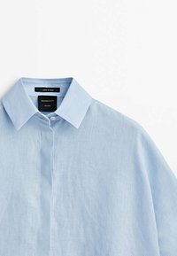 Chemise en lin bleu clair avec un col classique, dotée d'une fermeture à boutons et d'une texture subtile. L'étiquette indique "Fabriqué en Espagne" ainsi que le nom de la marque.