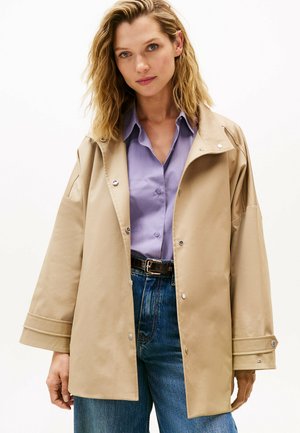 Beige oversized jas met drukknoopsluiting, met een hoge kraag en lange mouwen. Gdraagd over een lichtpaarse shirt en hooggeknipte blauwe jeans.