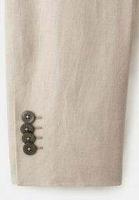Beige jakkesærm med fire lodret justerede mørkebrune knapper på manchet, tekstureret stof og synlig syning omkring knaphullerne.