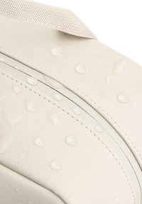 Sac imperméable de couleur crème avec une surface texturée, présentant des bords arrondis et des gouttes d'eau visibles, souligné par une couture contrastante.
