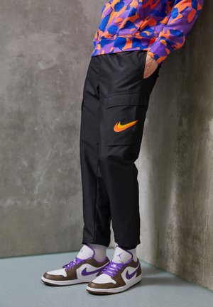 Person trägt schwarze Cargo-Hosen mit orangefarbenem Nike-Logo, ein lila-orange gemustertes Sweatshirt und weiße Turnschuhe mit lila Nike-Swoosh.