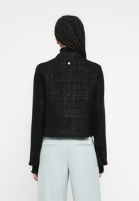 Jachetă neagră scurtă din material texturat, cu un model în carouri, mâneci lungi și un detaliu subtil cu nasture la gât.