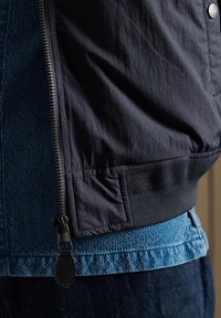Black nylon bomber jacket with elastic cuffs and hem, layered over a blue denim shirt. Features noticeable zipper and button details.