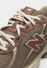 Καφέ και μπεζ αθλητικό παπούτσι New Balance με επάνω μέρος από πλέγμα, λευκά κορδόνια και μεγάλο λογότυπο "N" στο πλάι, που δείχνει λεπτομερή υφή και σχεδιασμό.
