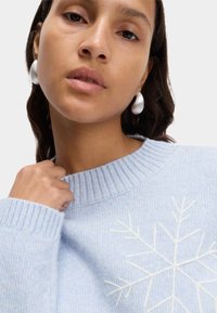 Jasnoniebieski sweter z dzianiny z prążkowanym dekoltem i białym haftem w śnieżynki z przodu; modelka nosi duże, perłowe kolczyki.