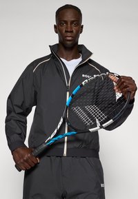 Melna jaku ar rāvējslēdzēju un baltām un zilām akcentiem, ar elastīgām piedurknēm. Modelis tur melnu un zilu tenisa racketu ar strukturētām stīgām.