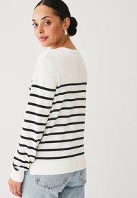 Maglione bianco a righe con strisce orizzontali nere, colletto rotondo, polsini e orlo a costine, abbinato a jeans azzurri chiari. Materiale in maglia morbida.