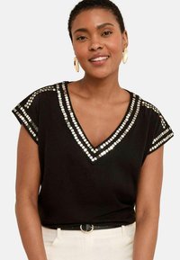 Haut noir à manches courtes avec un col en V, orné de sequins circulaires argentés le long de l'encolure et des épaules. Tissu lisse et coupe décontractée.