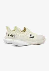Sneakers Lacoste con una parte superiore in rete color crema, suola bianca texturizzata e dettagli del logo. Accenti gialli sul tallone e sulla linguetta. Ammortizzazione LSRFOAM.