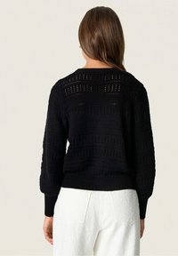 Žena s dlouhými hnědými vlasy, která má na sobě černý pletený svetřík s dlouhým rukávem a bílé texturované kalhoty, tváří k nám zády před jednobarevným pozadím.