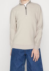 Uomo che indossa un pullover beige a zip parziale e jeans di denim blu larghi, in piedi contro uno sfondo bianco semplice.