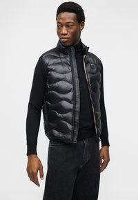 Άνδρας που φοράει μαύρο αμάνικο καπιτονέ γιλέκο πάνω από μαύρο μακρυμάνικο πουκάμισο και σκούρο τζιν, κοιτάζοντας προς τα αριστερά του, με φόντο μια μονόχρωμη επιφάνεια.