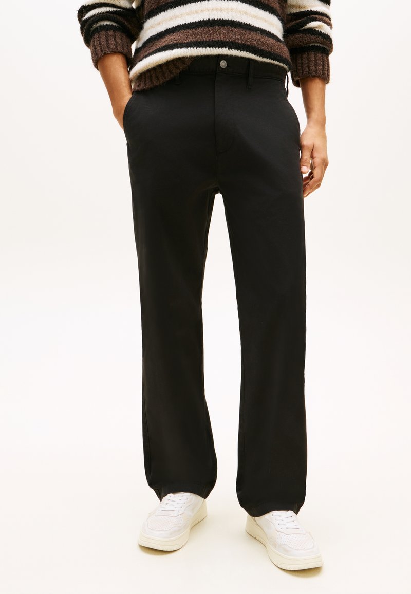 Fekete egyenes szárú nadrágot, barna-fehér csíkos pulóvert és fehér tornacipőt viselő személy, részben a zsebében tartott kézzel áll.