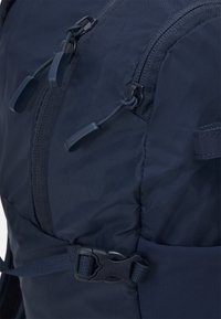 Tamsiai mėlynas kuprinės modelis su tekstūruotu audiniu, dviem užtrauktukais su kilpomis ir šonine sagtimi tvirtesniam užsegimui.