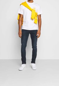 Vit t-shirt med gult solstrålelogotyp, kombinerad med mörkblå jeans och vita sneakers. Gult sweatshirt hängande över axlarna.