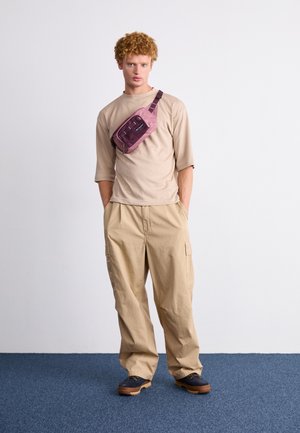 Junger Mann mit lockigen Haaren steht auf blauem Teppich, trägt ein beiges Hemd, khakifarbene Cargohose, dunkle Schuhe und eine mauvefarbene Umhängetasche.