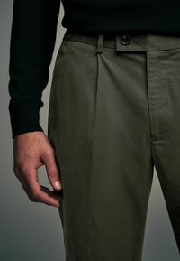 Pantalons khaki avec un design ajusté, plis à l'avant et une fermeture à bouton, associés à un pull sombre. Tissu texturé.