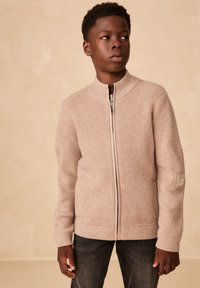 Suéter beige de punto con cuello alto y cremallera de largo completo. Presenta textura acanalada y detalle de parche en la manga izquierda. Lleva puesto sobre jeans oscuros.