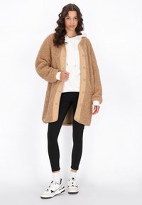 Cappotto in pile beige con fodera beige, caratterizzato da grandi bottoni e comode maniche oversize. Indossato sopra un felpa bianca e leggings neri.