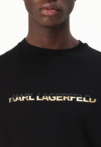 Melna T-kreklā ar īsām piedurknēm, uz krūtīm ir izteikts zelta un melns logotips. Izgatavots no mīksta kokvilnas auduma ar apaļu kakla izgriezumu.