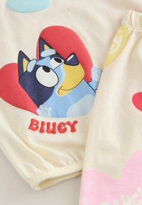 Pantaloni da pigiama chiari con un grande cane cartoon in blu e giallo, su uno sfondo rosso a forma di cuore e la parola "BLUEY" in rosso.