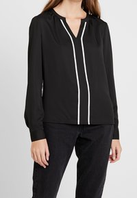 Svart långärmad blus med V-ringning, med vit vertikal piping längs fram. Slätt material, avslappnad passform, subtila sammankopplingar vid axlarna.