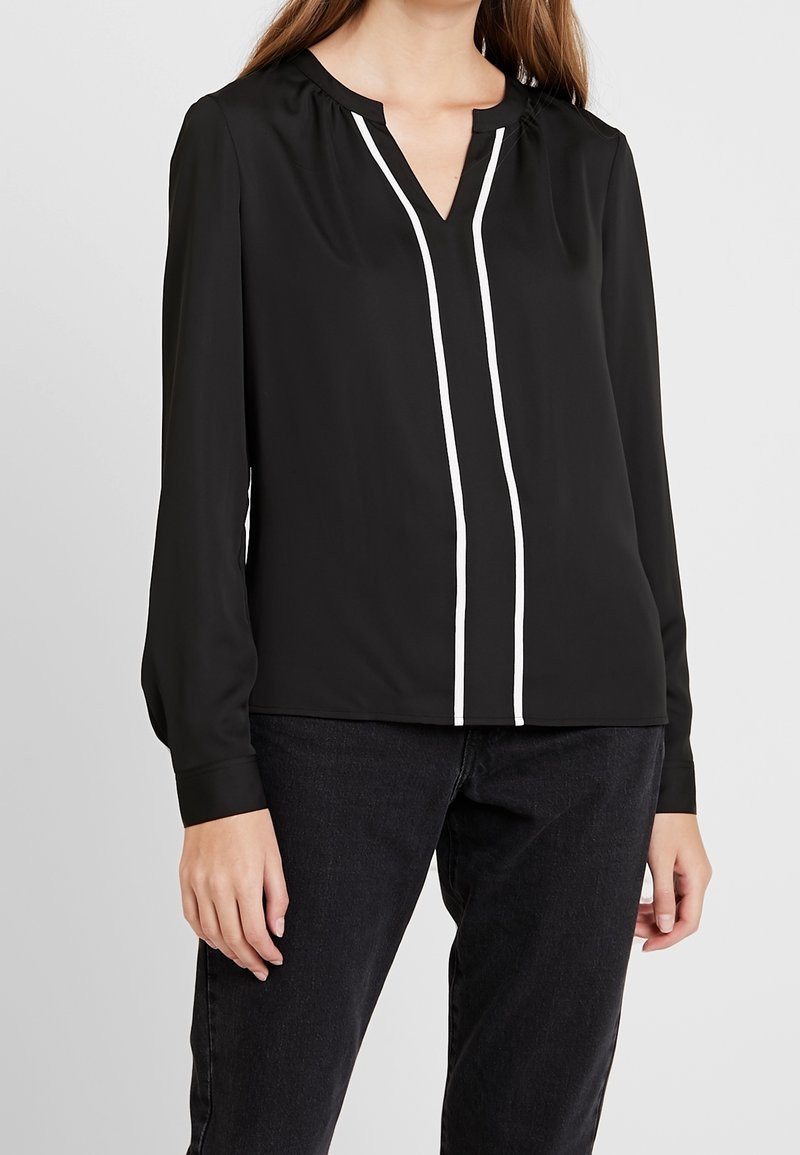 Svart långärmad blus med V-ringning, med vit vertikal piping längs fram. Slätt material, avslappnad passform, subtila sammankopplingar vid axlarna.