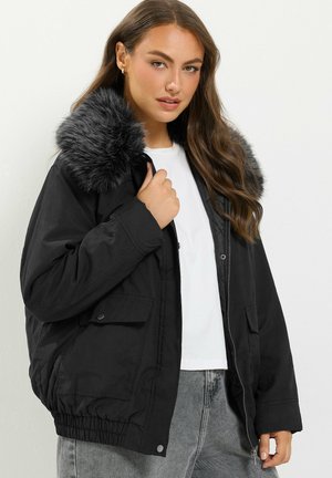 Femme portant une veste noire avec un grand col en fausse fourrure grise, chemise blanche et jean gris, posant avec une main tenant le col de la veste.
