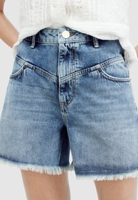 Pantalones cortos de mezclilla azul claro con un dobladillo desflecado, cintura de tiro medio y diseño de cinco bolsillos. Presenta costuras en contraste y un cierre de botón metálico.