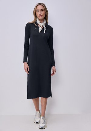 Kvinde iført en sort midikjole med lange ærmer, hvidt mønstret halstørklæde og hvide sneakers, stående mod en ensfarvet lys baggrund.