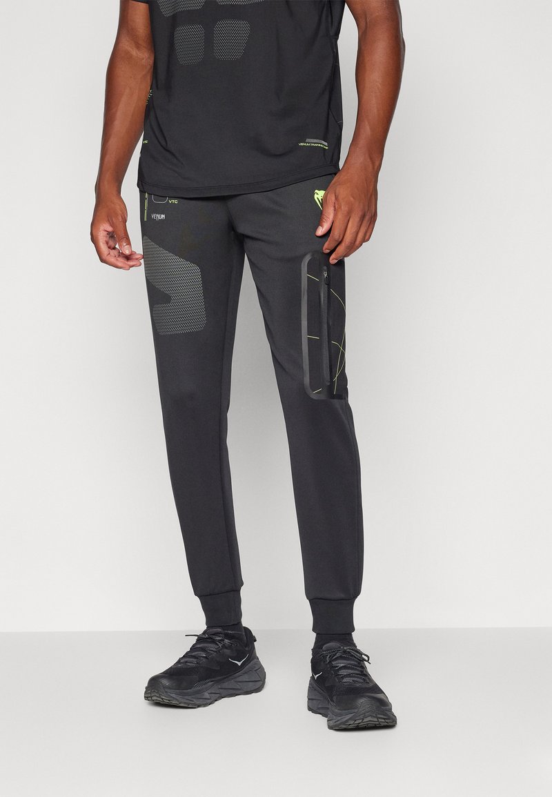 Sorte atletiske bukser med slim fit, der har mesh-mønstre, lynlåslommer og grøn akcentstikning langs siderne.
