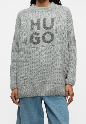 Person trägt einen locker sitzenden grauen Strickpullover mit großen schwarzen Buchstaben "HUGO", kombiniert mit hellblauen Jeans.