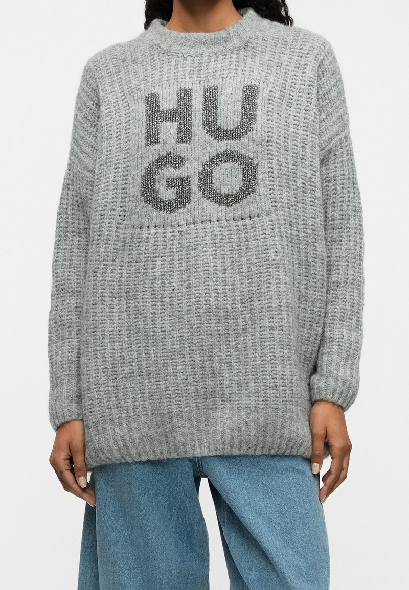 Personne portant un pull en maille gris ample avec "HUGO" inscrit en grandes lettres noires, associé à un jean en denim bleu clair.