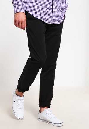Mann trägt schwarze Hose, weiße Sneakers und ein lila kariertes Hemd, mit einer Hand in der Hosentasche, geht auf einem einfarbigen hellen Hintergrund.