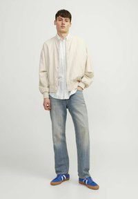 Cazadora bomber beige claro, camisa de rayas blanca y azul, jeans azul claro y zapatillas azules con rayas blancas. Diseño simple y casual.