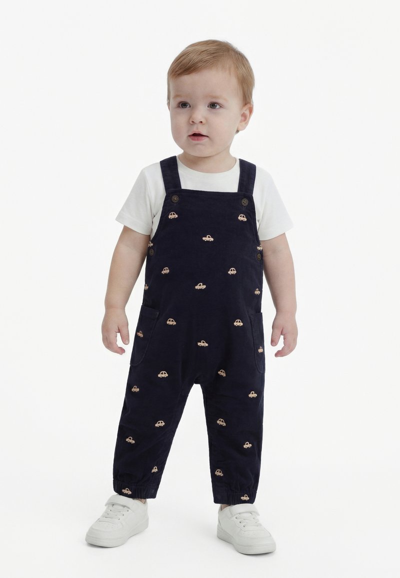 Tout-petit debout portant une salopette noire avec des petits imprimés de voitures, un t-shirt blanc à manches courtes, et des baskets blanches sur un fond uni.