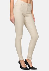 Jeans skinny beiges en tissu lisse et extensible. Dotés d'une taille haute et d'un design classique à cinq poches. Associés à des escarpins noirs à bout pointu.