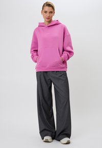 Rozā kapucēta sporta krekls ar priekšējo kabatu, apvienots ar pelēkām svītrainām plaša griezuma biksēm un gaišas krāsas sporta apaviem. Vienkārša dizaina; maiga tekstūra.