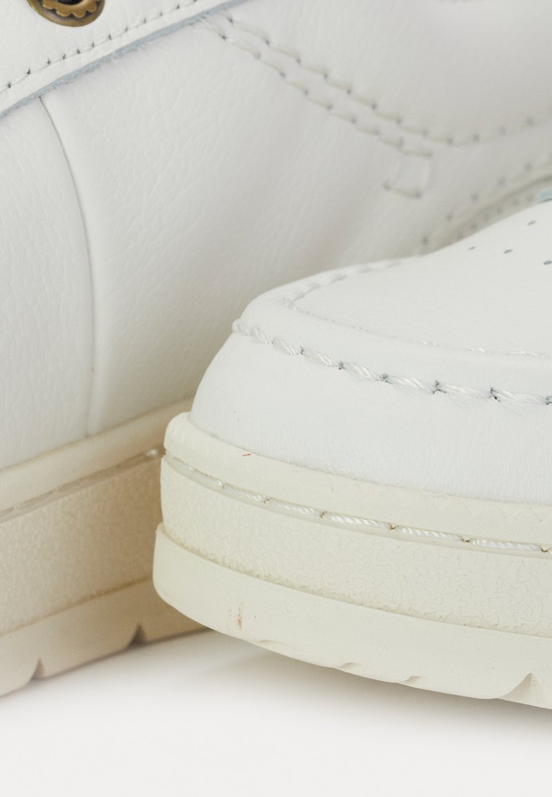 Baskets blanches en matériau synthétique lisse. Les caractéristiques incluent des détails de couture, des perforations et une semelle en caoutchouc texturé.