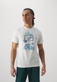 Camiseta de algodón blanca que presenta un gráfico con un aro de baloncesto, hojas de palmera y "JUST DO IT" en tonos azules y verdes. Manga corta.