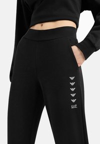 Pantaloni sportivi neri in tessuto morbido e elasticizzato, con vita alta, tasche laterali e un motivo con logo lungo il lato sinistro.